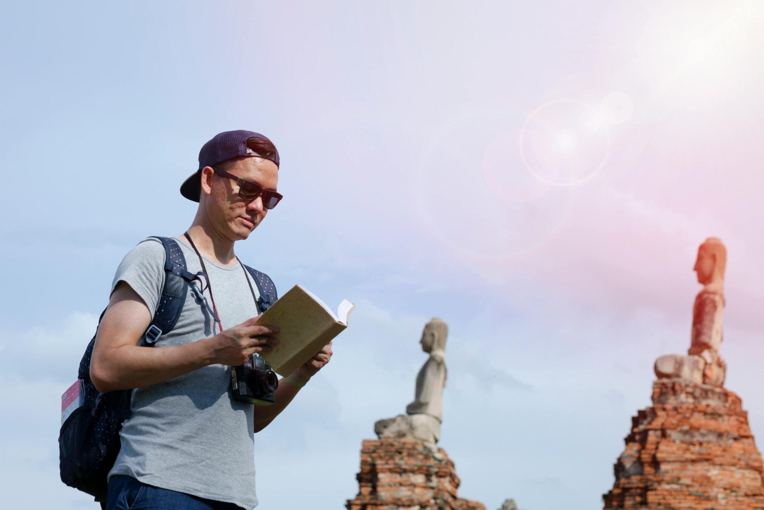 A tourist with sunglasses reads a book near ancient statues under daylight.